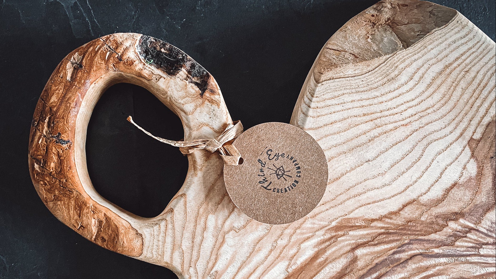 A rustic wooden serving board with a thick, rounded handle and a natural live edge, featuring prominent wavy grain patterns in light and dark brown tones. A round kraft paper tag is attached to the handle with twine.