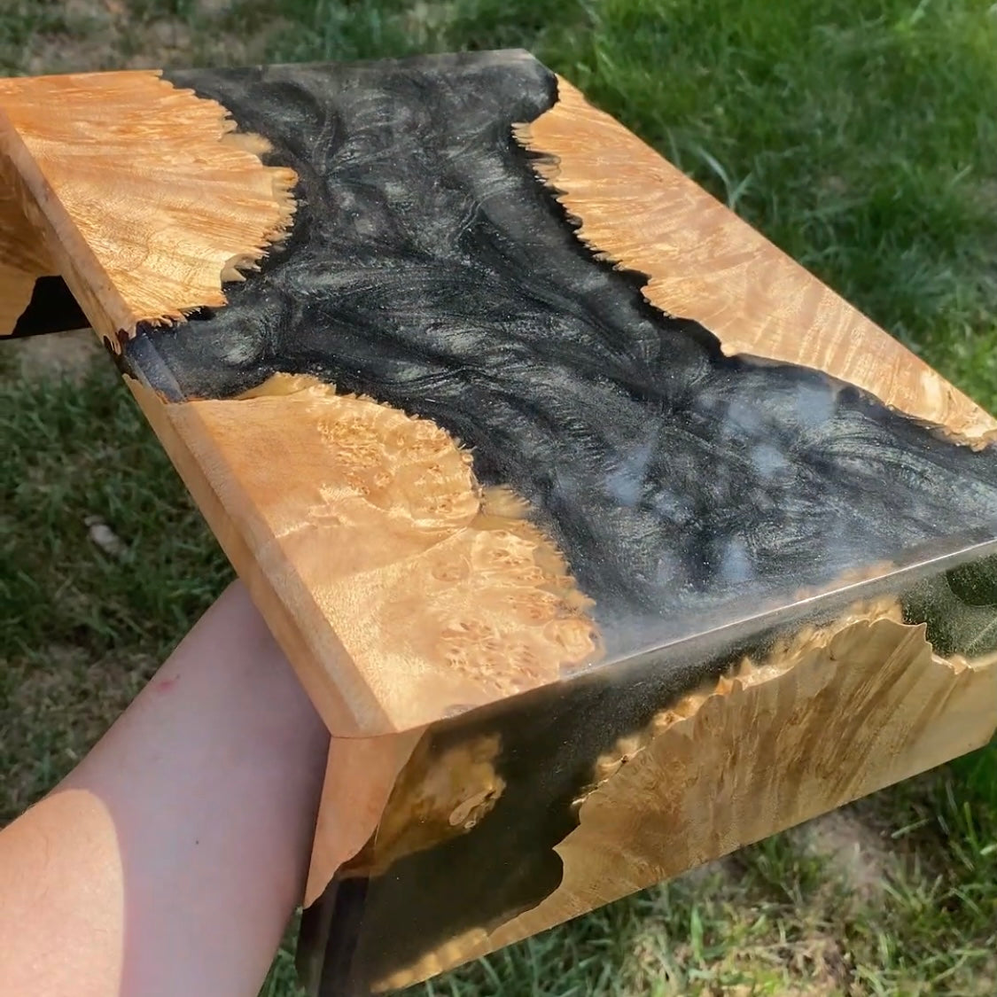 A close-up of a monitor stand made of maple burl with a dramatic black resin inlay flowing through a natural light tan wood grain, held by a person's arm outdoors on grass.