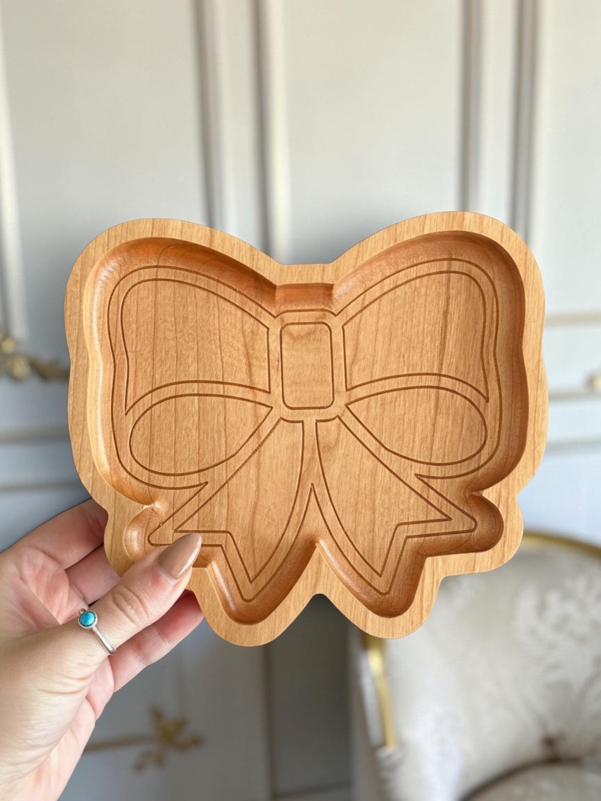 A hand holding a small wooden tray shaped like a bow, showing the natural light wood grain and carved interior details, with a softly blurred indoor background.
