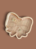A bow-shaped wooden jewelry tray, shown on a solid brown background with two small gold rings placed inside.
