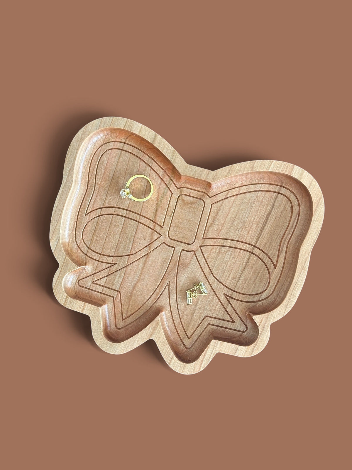 A bow-shaped wooden jewelry tray, shown on a solid brown background with two small gold rings placed inside.