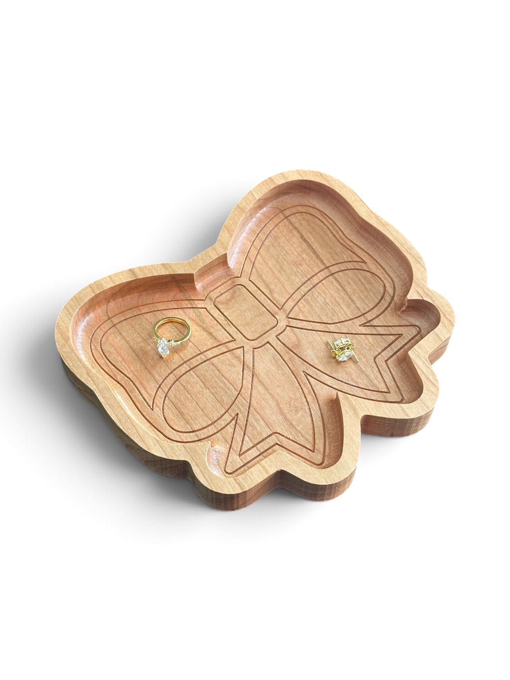 A bow-shaped wooden ring tray with a natural light wood finish and engraved details, holding two small rings, on a clean white background.