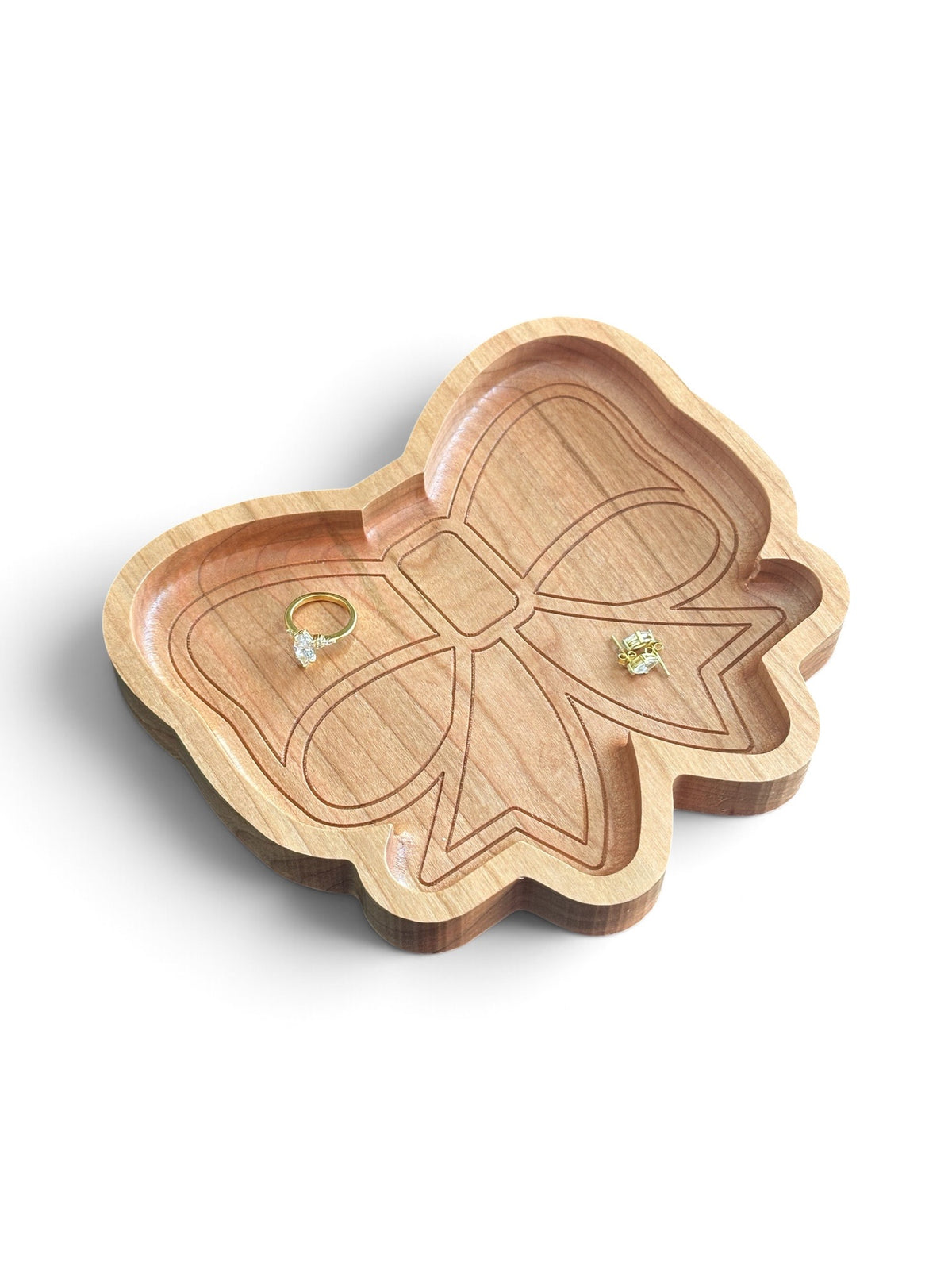 A bow-shaped wooden ring tray with a natural light wood finish and engraved details, holding two small rings, on a clean white background.