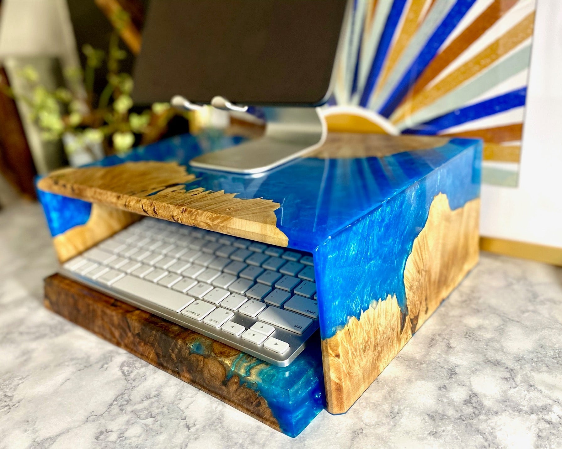 A desktop computer monitor stand made from burl and a blue river wood grain resin finish, holding a keyboard and monitor stand on a marble surface.