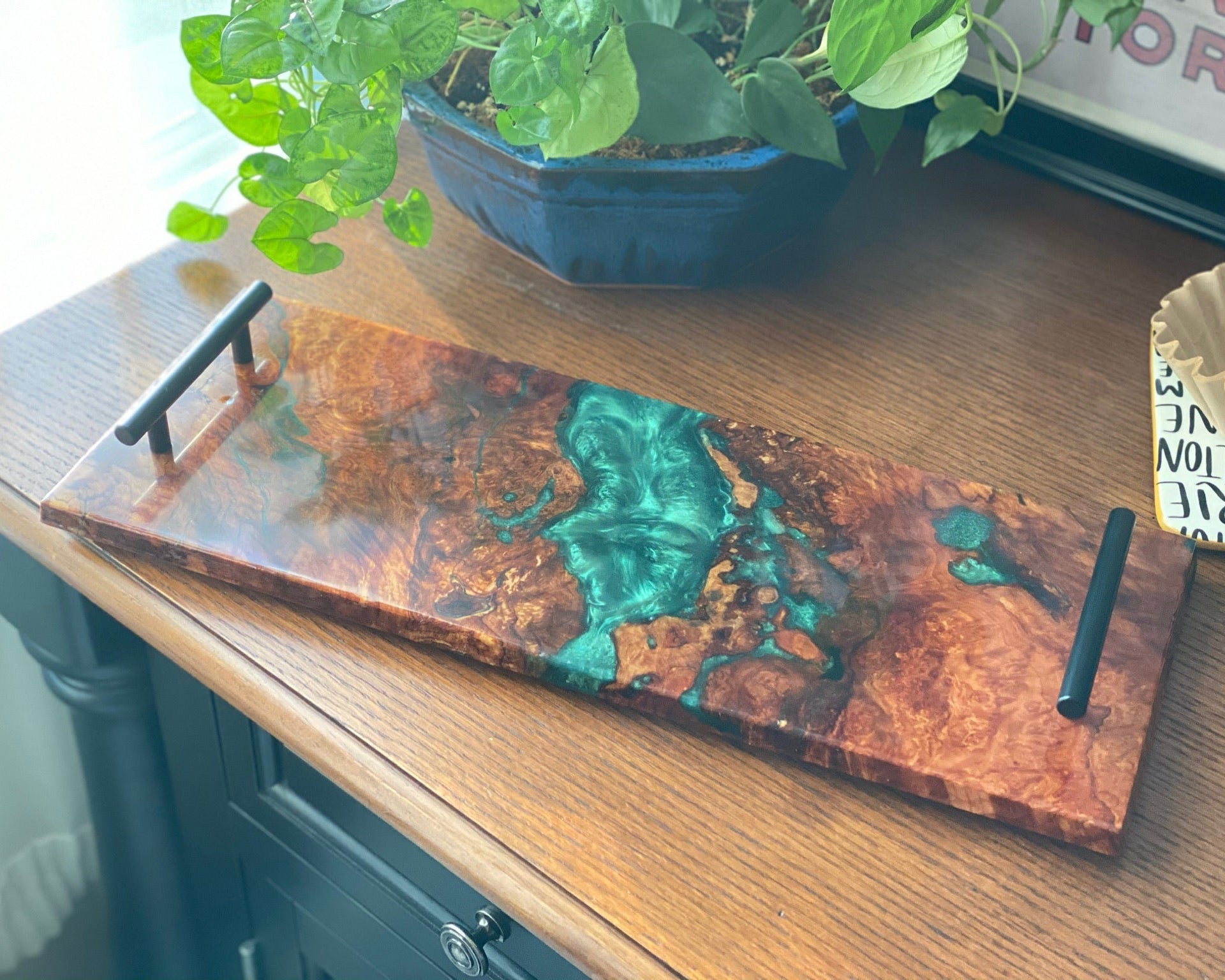 A rectangular wooden serving tray with a glossy finish, featuring a swirling pattern of copper-brown wood grain and vibrant green resin inlay. It has two black metal handles, one at each end, and is displayed on a wooden table with a potted plant in the background.