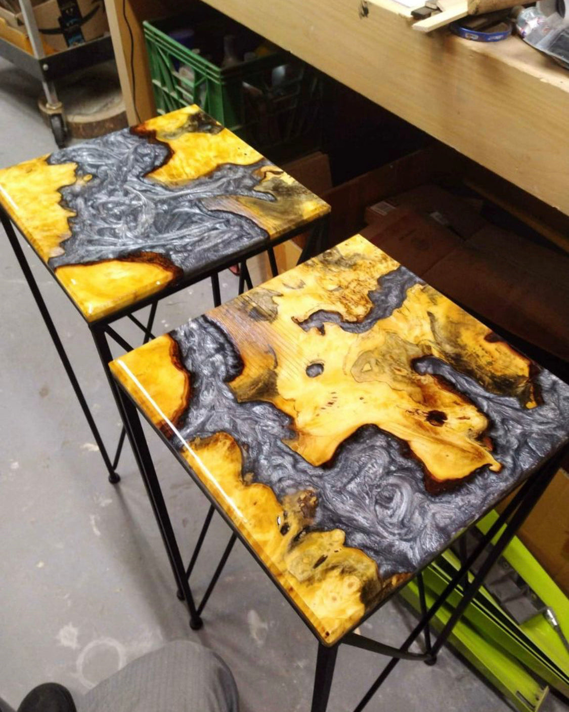 Two square hairpin table stands with wood burl and glossy resin tops featuring a natural wood grain pattern in yellow, brown, and dark gray, supported by black metal legs in a workshop setting.