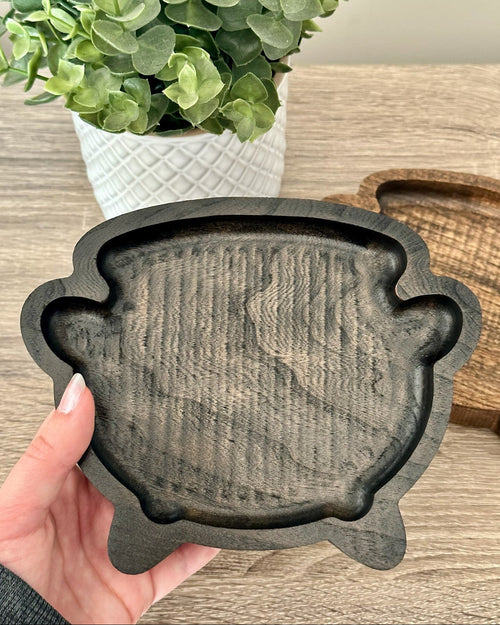 A hand holding a black cauldron wooden tray set against a rustic wooden tabletop with a potted green plant in the background.