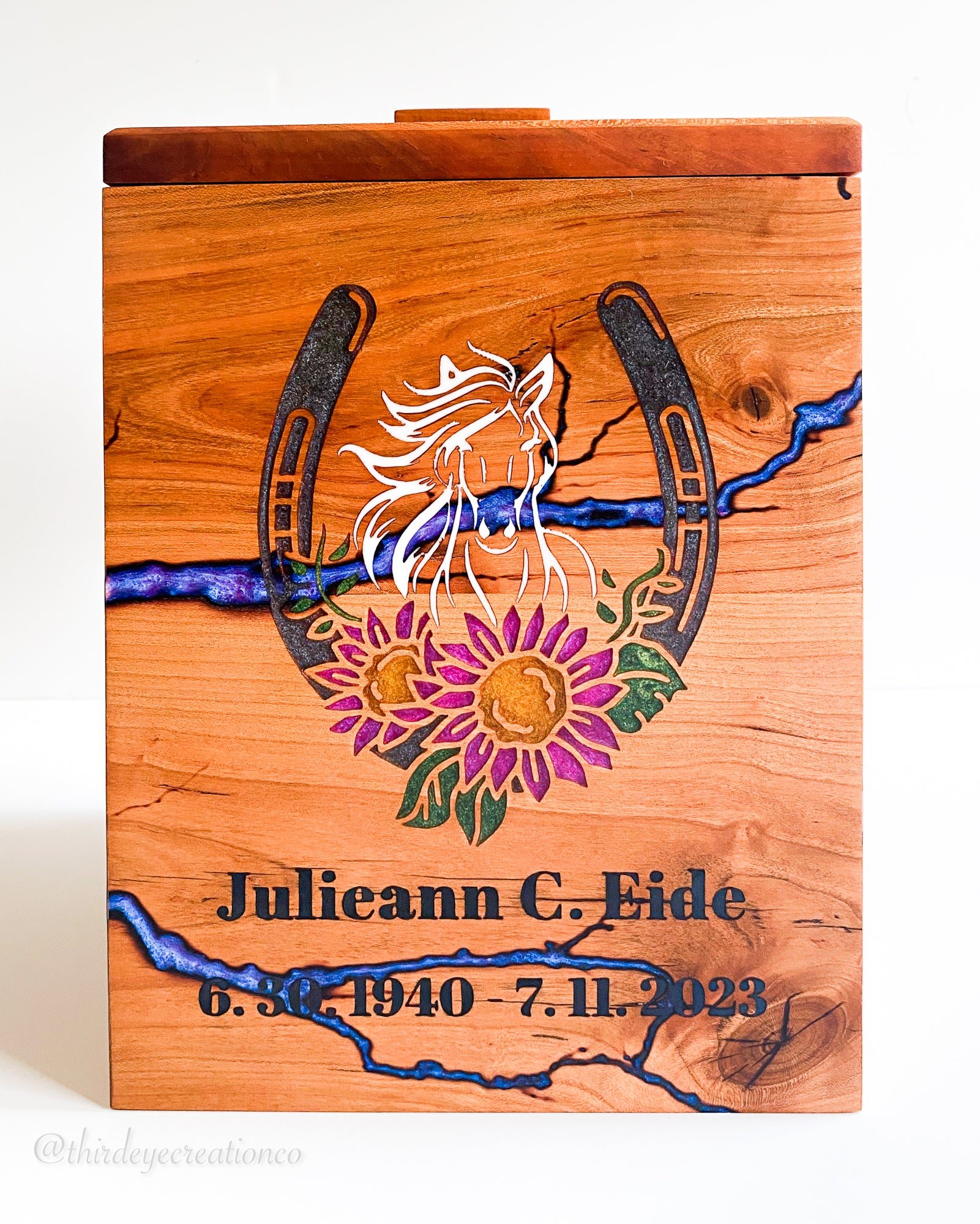A personalized wooden urn box with a lid, featuring a resin inlay horseshoe with a white horse silhouette and purple, pink, and green flowers and purple fractal burns in the background. The box is engraved with the name 'Julieann C. Eide’. 