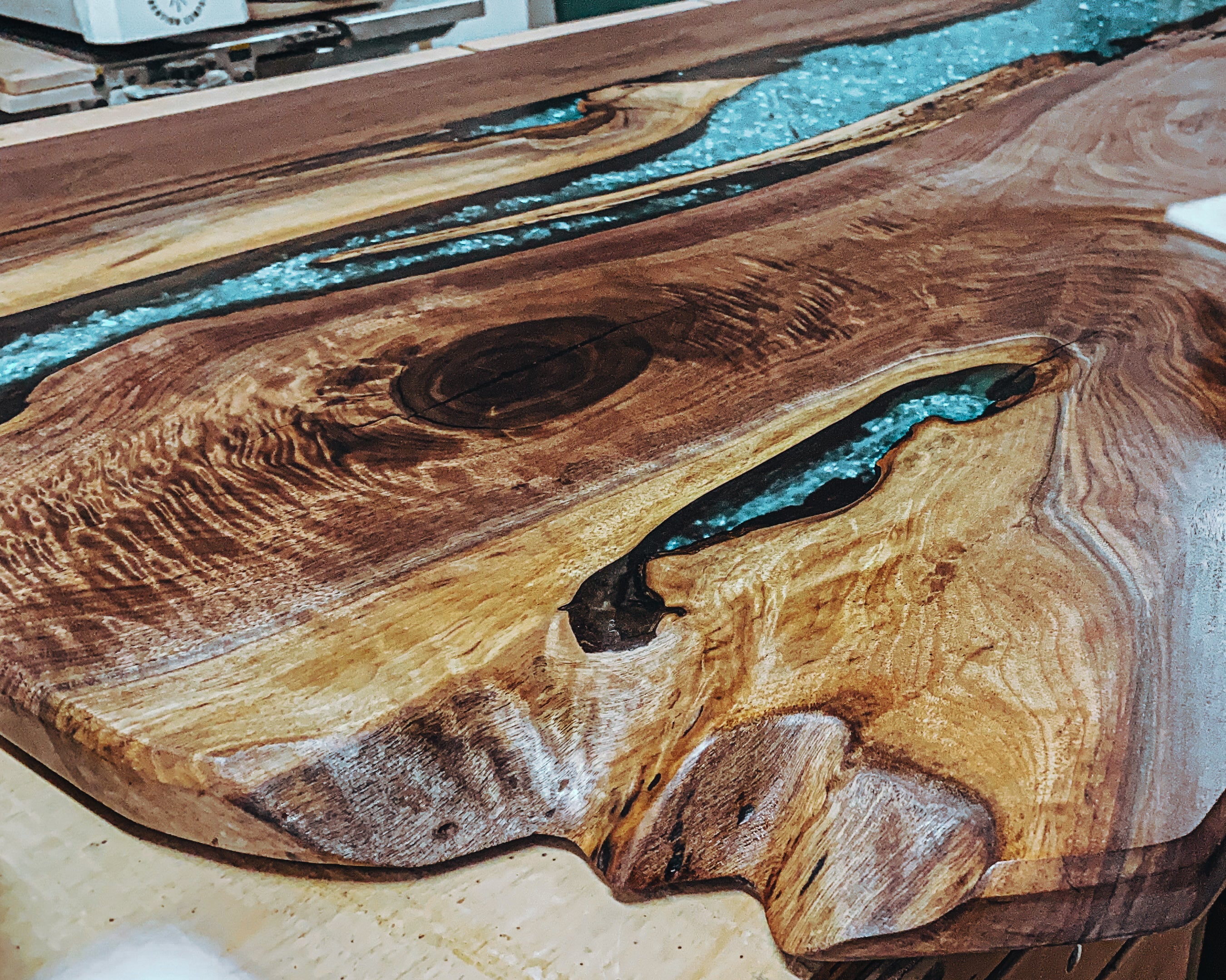 A large live-edge wooden tabletop with a turquoise resin river running through the center, shown in a workshop with a yellow DeWalt orbital sander working on the wood surface.