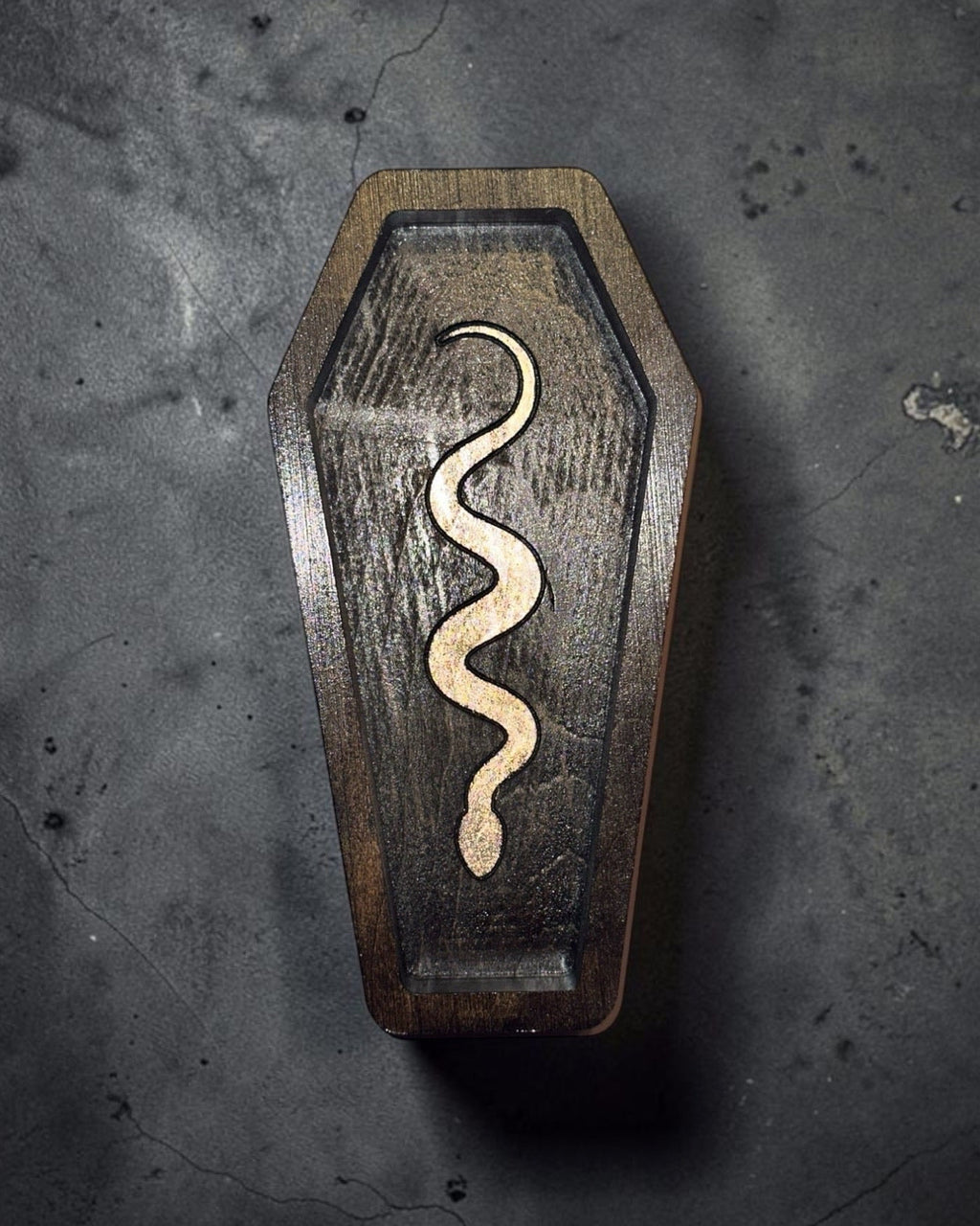 A coffin-shaped wooden box with a carved snake design on the front, photographed from above on a dark textured surface.