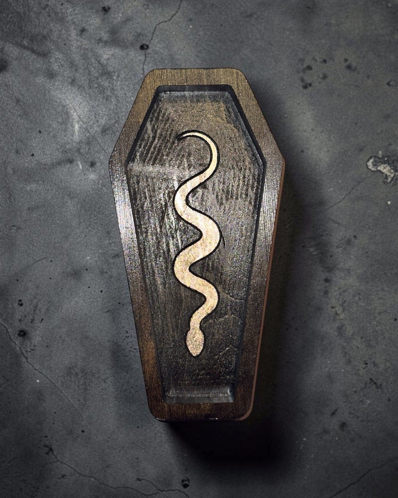 A coffin-shaped wooden box with a carved snake design on the front, photographed from above on a dark textured surface.