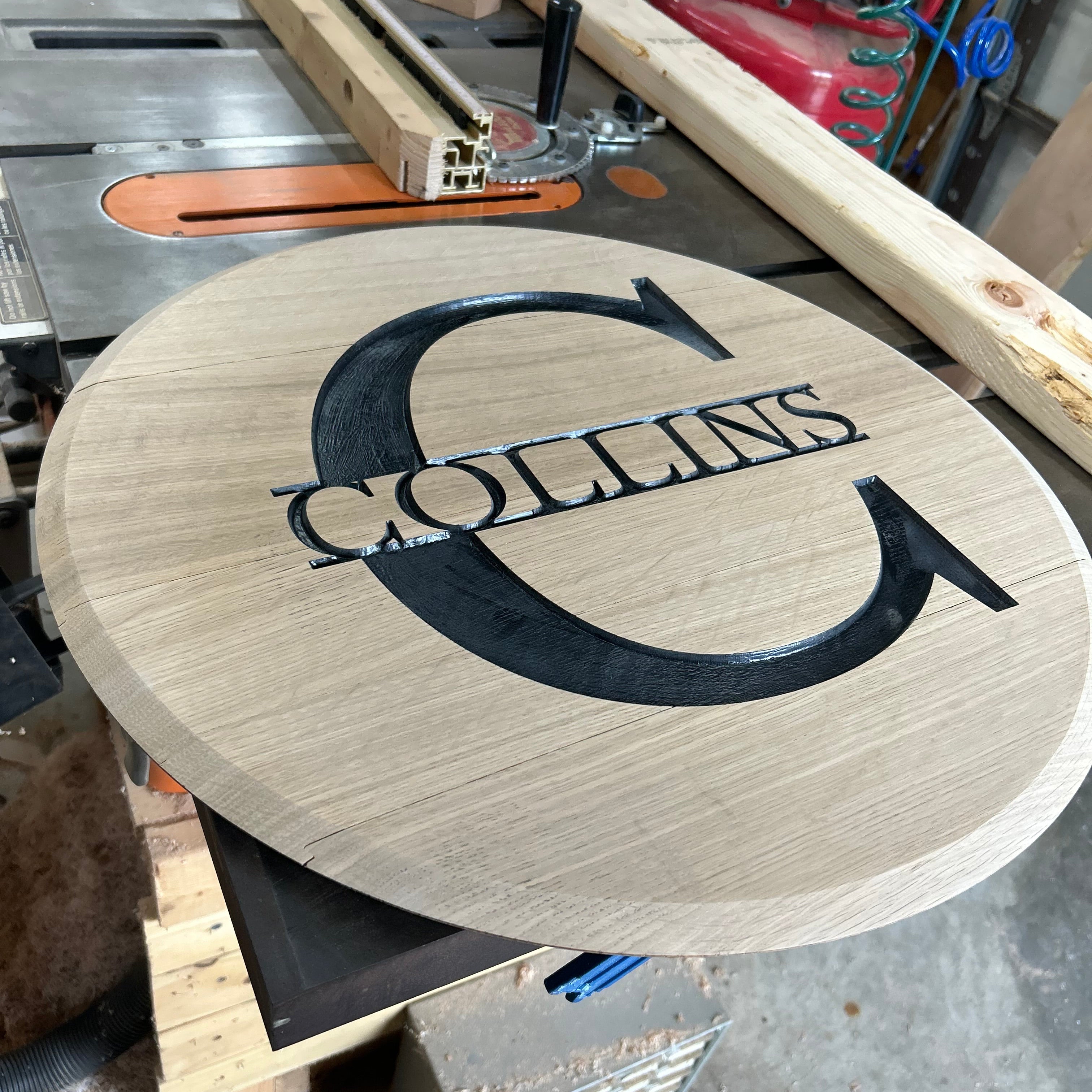 A round wooden barrel lid with a natural oak finish, featuring a black engraved logo of a stylized 'C' and the name 'COLLINS' across the center, resting on a workbench in a woodworking shop.