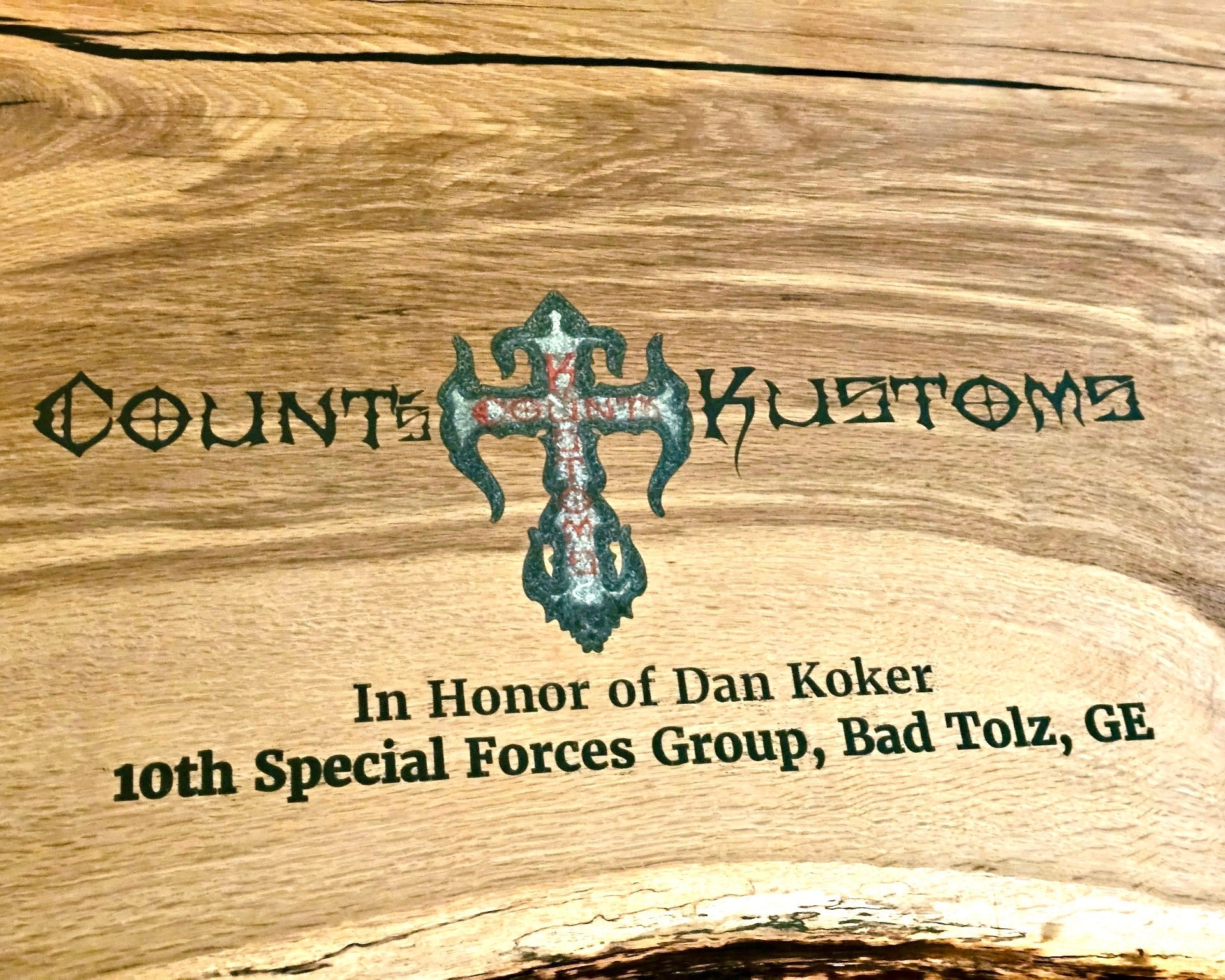 A close-up of a table or sign an inlay resin text reading 'Count Kustoms' and a central ornate cross emblem, plus the inscription 'In Honor of Dan Koker' and '10th Special Forces Group, Bad Tolz, GE'.