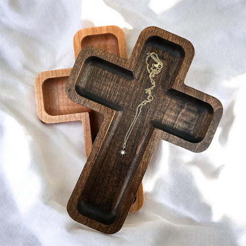 A dark brown wooden cross-shaped tray holding a delicate gold necklace with a star pendant, a lighter natural wood cross trays stacked behind it on a white fabric background.