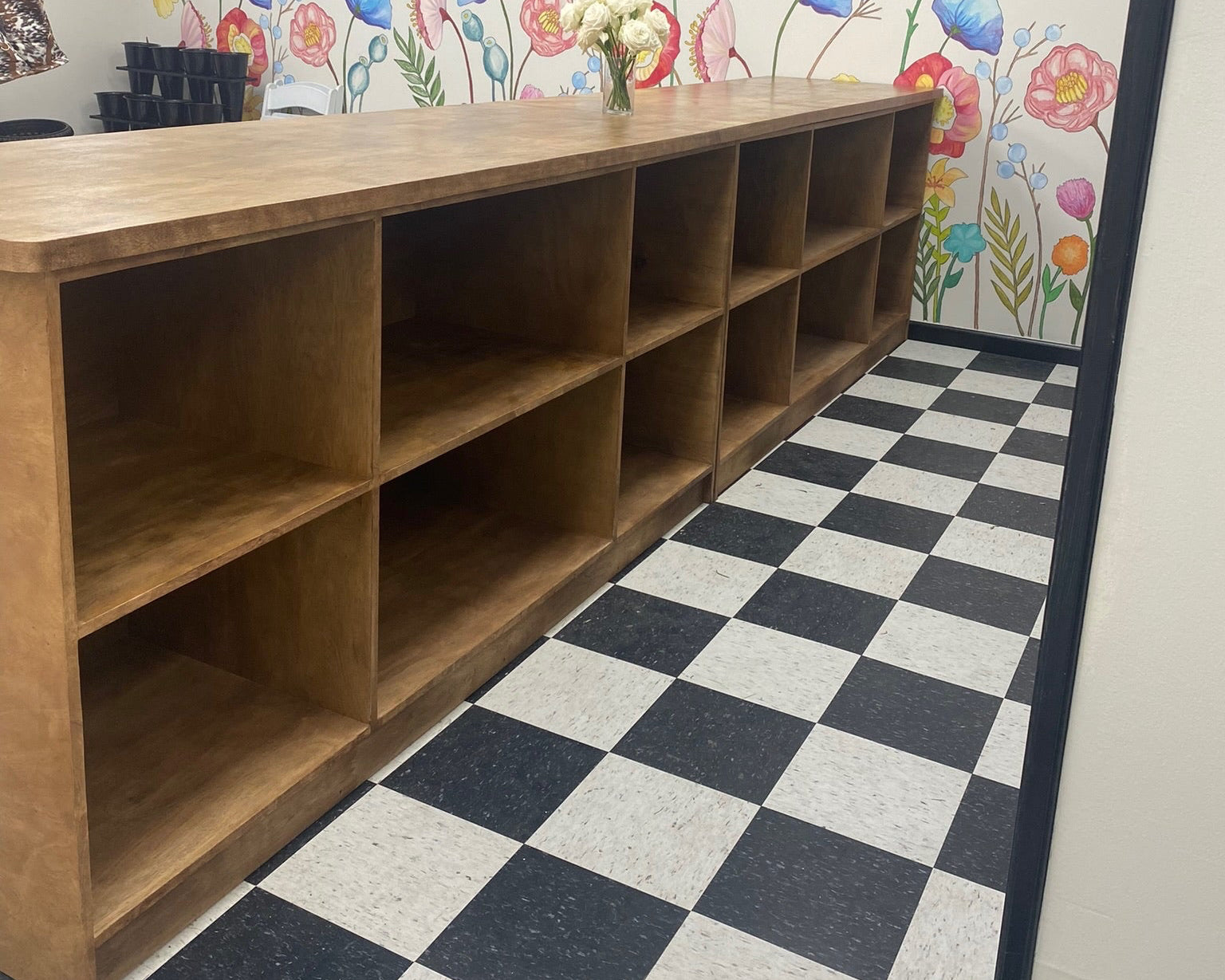 Wooden checkout shelving unit in a floral shop with a checkered floor and floral wall art.
