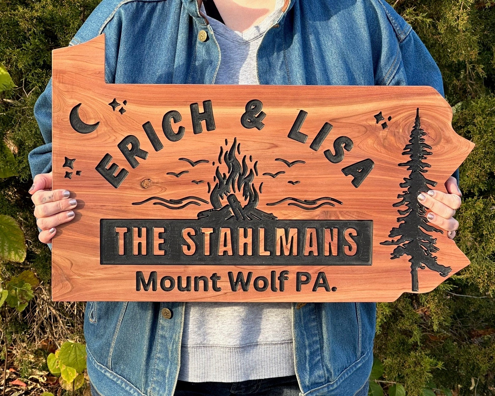 A person outdoors holding a custom wooden sign shaped like a mountain range. The sign is engraved with the names 'ERICH & LISA', 'THE STAHLMANS', and 'Mount Wolf PA.