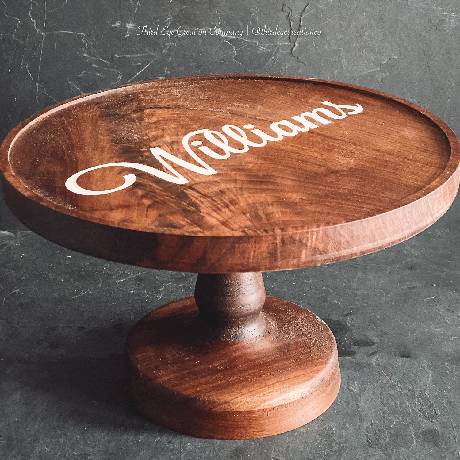 A round wooden pedestal table or cake stand with a turned wood base, featuring the name 'Williams' in white resin inlay script on the top surface, set against a dark textured gray background.