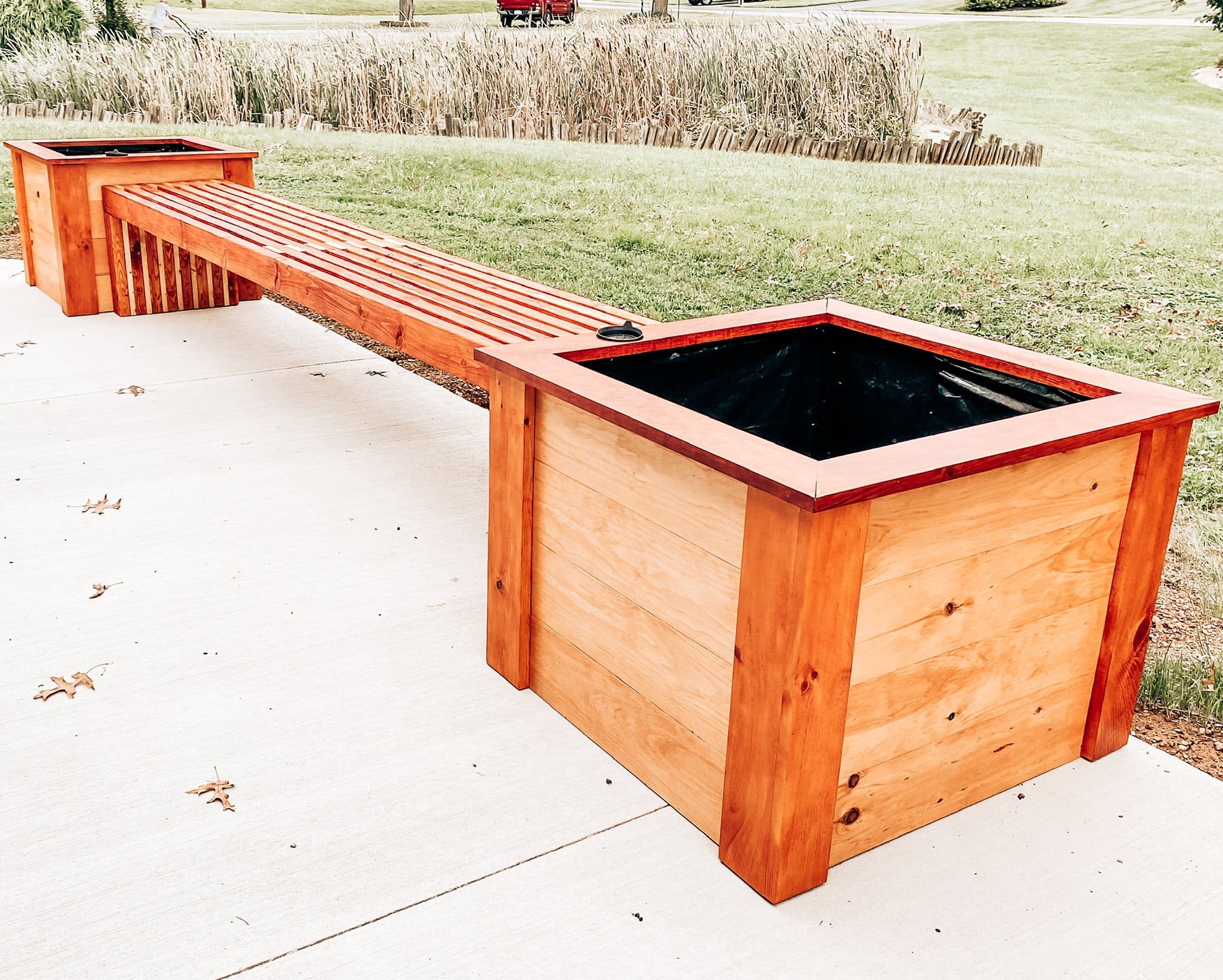 A long, rectangular outdoor wooden bench with integrated planter boxes, shown on a concrete patio with grass and reeds in the background. A watermark at the bottom reads 'Third Eye Creation Company | @thirdeyecreationco'.