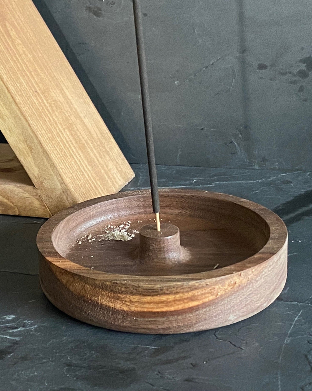 A round wooden incense holder with a central stand and a single incense stick inserted, placed on a dark stone surface. A light wood display block and a potted green plant are visible in the background.