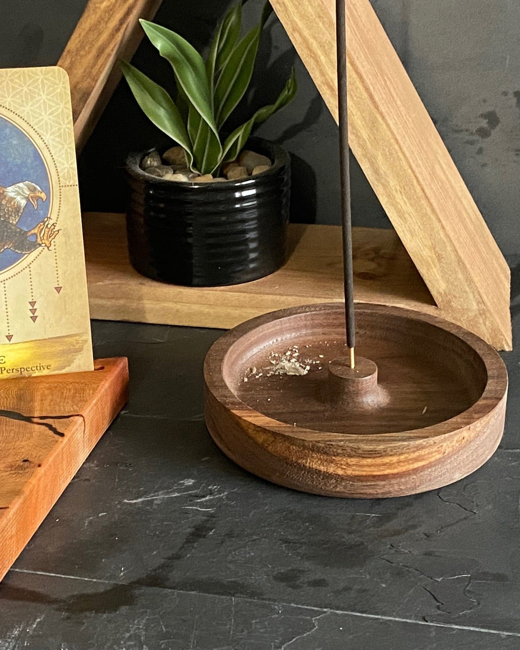 A tarot card set featuring an 'Eagle' card with a wooden stand, displayed alongside a wooden incense holder with a burning incense stick and a small potted plant in the background.