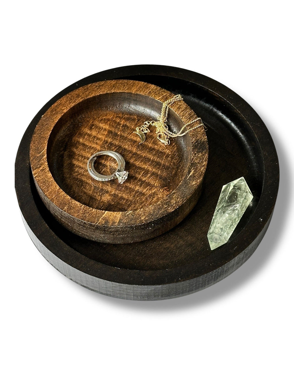 Two round wooden jewelry trays, one small dark brown tray nested inside a larger black tray, styled with a silver ring, gold necklace, and a clear crystal on a white background.