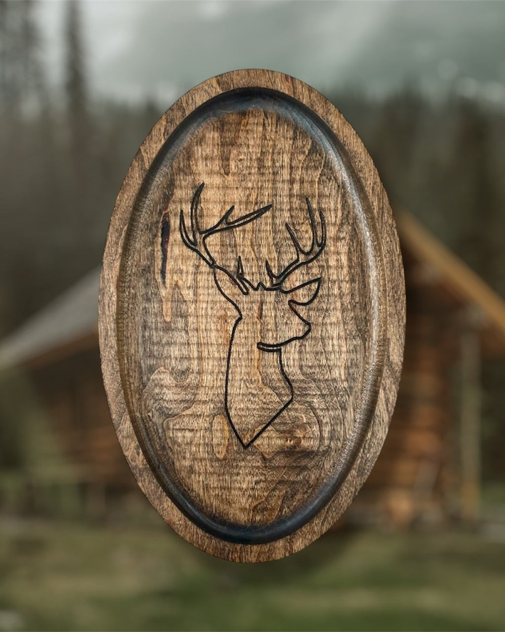 An oval wooden tray with a carved deer head profile, shown against a blurred outdoor background of a cabin and trees.