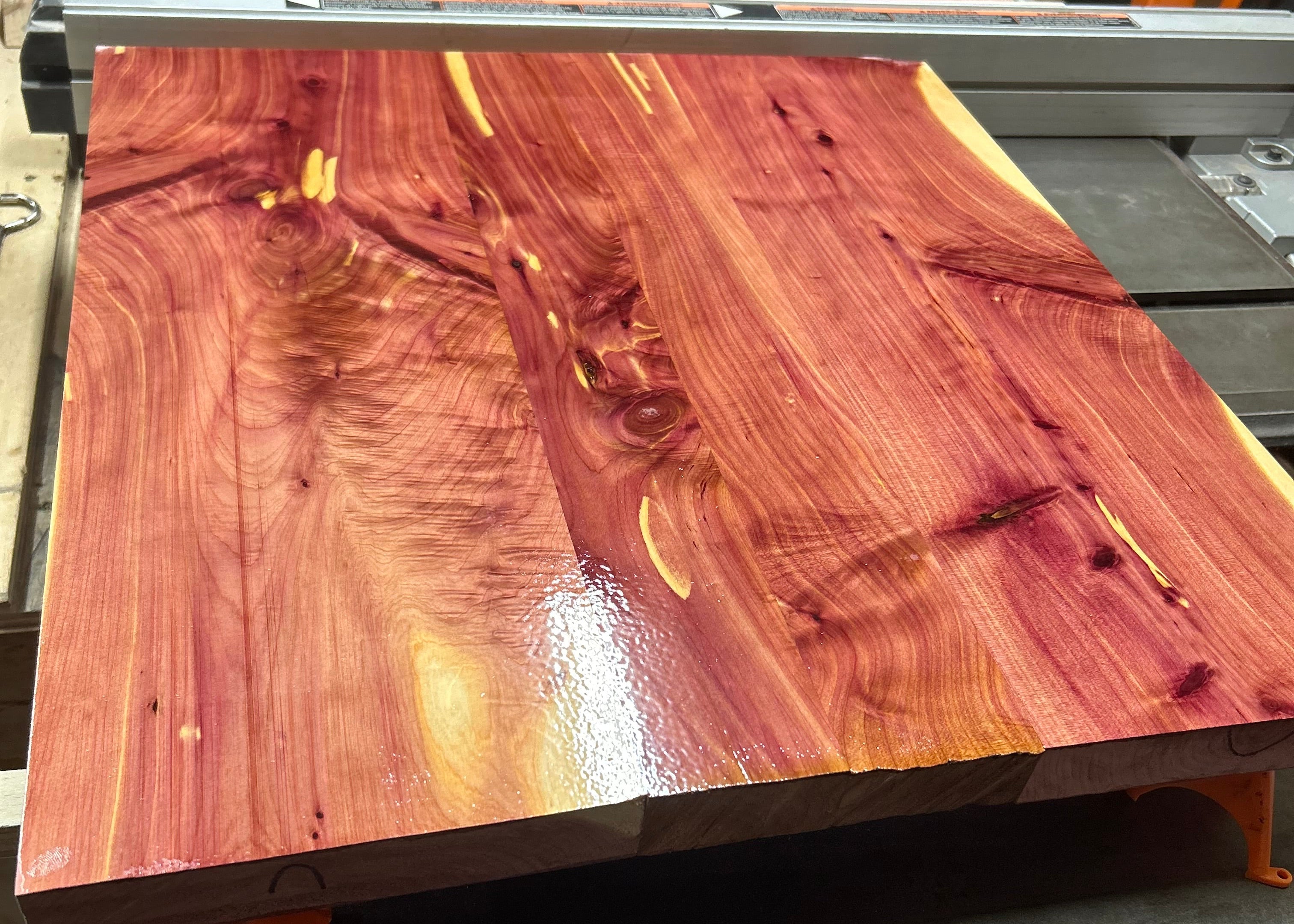 A large, glossy live-edge cedar wood slab with rich reddish-brown and golden grain patterns, resting on a metal table saw in a workshop setting.