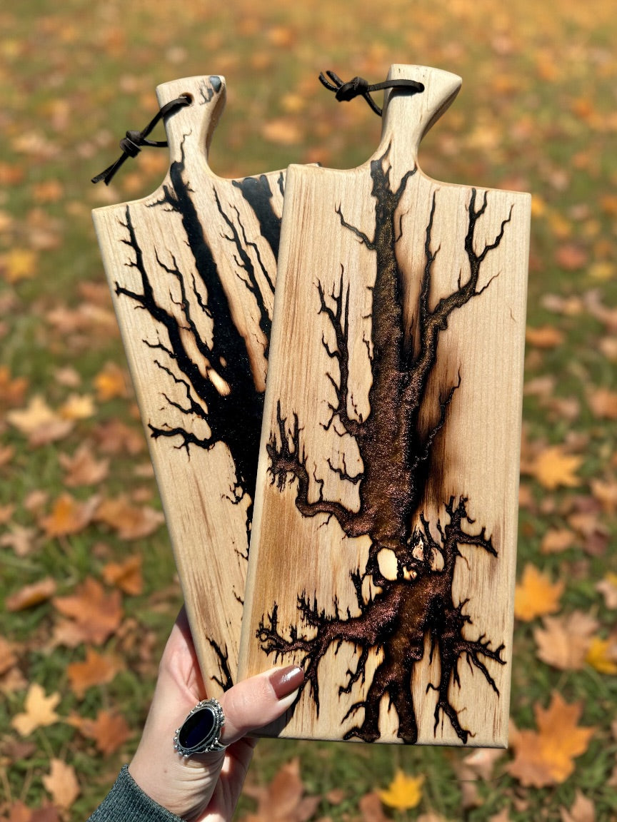 Two rectangular wooden cutting boards with rounded handles, held by a hand against a background of grass and fallen autumn leaves. The boards feature intricate black and brown branch engravings.