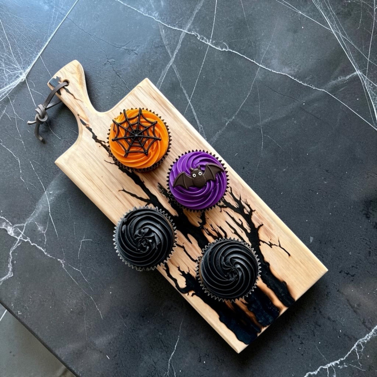 A Halloween-themed wooden cutting board with a handle, decorated with black spiderwebs and tree branches, holding four decorated cupcakes on a dark gray marble surface.
