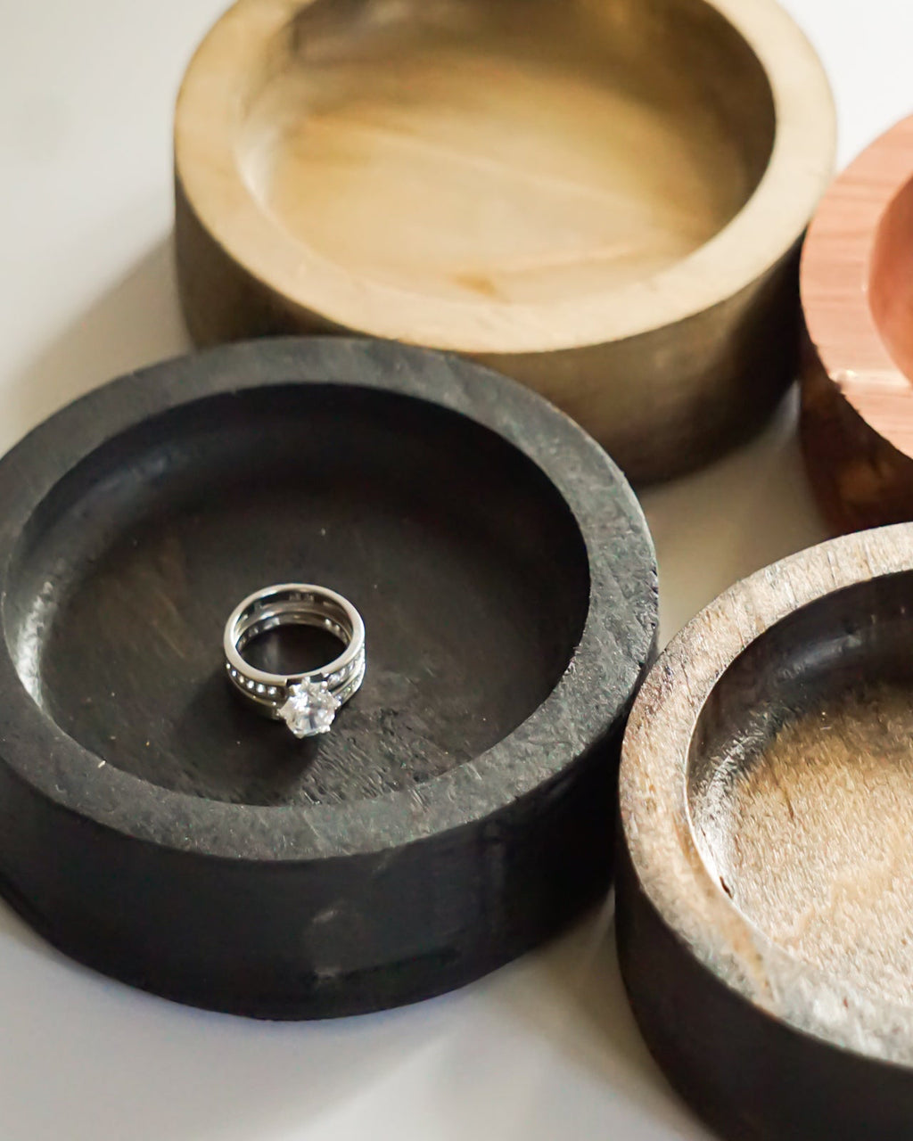 Four round wooden ring dishes  in different wood tones, arranged on a light background. One dish holds a silver ring, and the light brown dish holds a clear crystal.