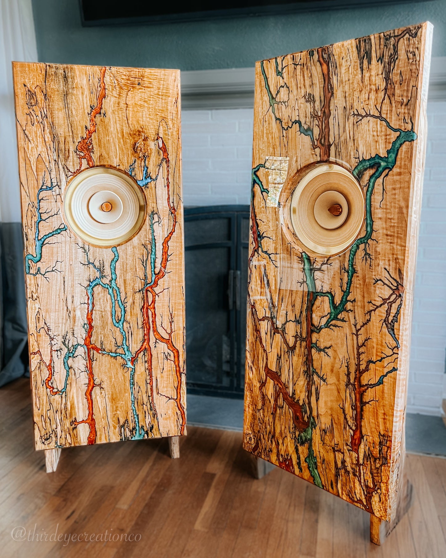 Two tall, rectangular wooden maple speaker stand with exposed wood grain and colorful fractal burned inlay patterns, standing on a wooden floor in front of a fireplace.