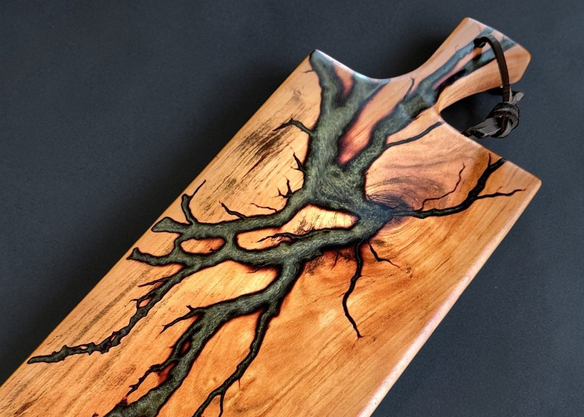 A rectangular wooden serving board with a natural wood grain finish and a prominent dark resin fractal burn tree branch inlay, shown on a dark textured background.