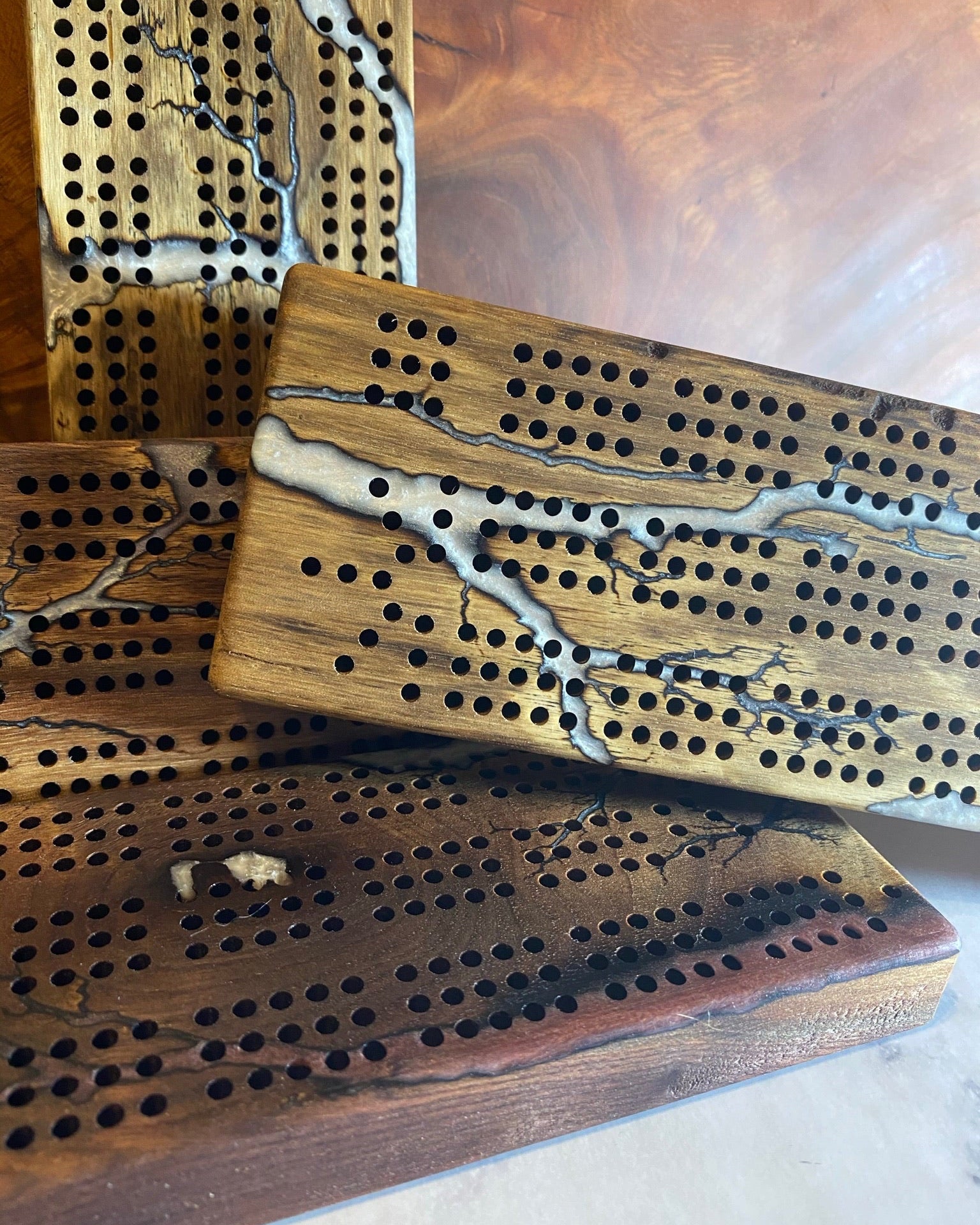Four wooden cribbage boards with engraved holes and natural wood grain, featuring white resin inlay with dark branch-like patterns, stacked on a wooden surface.