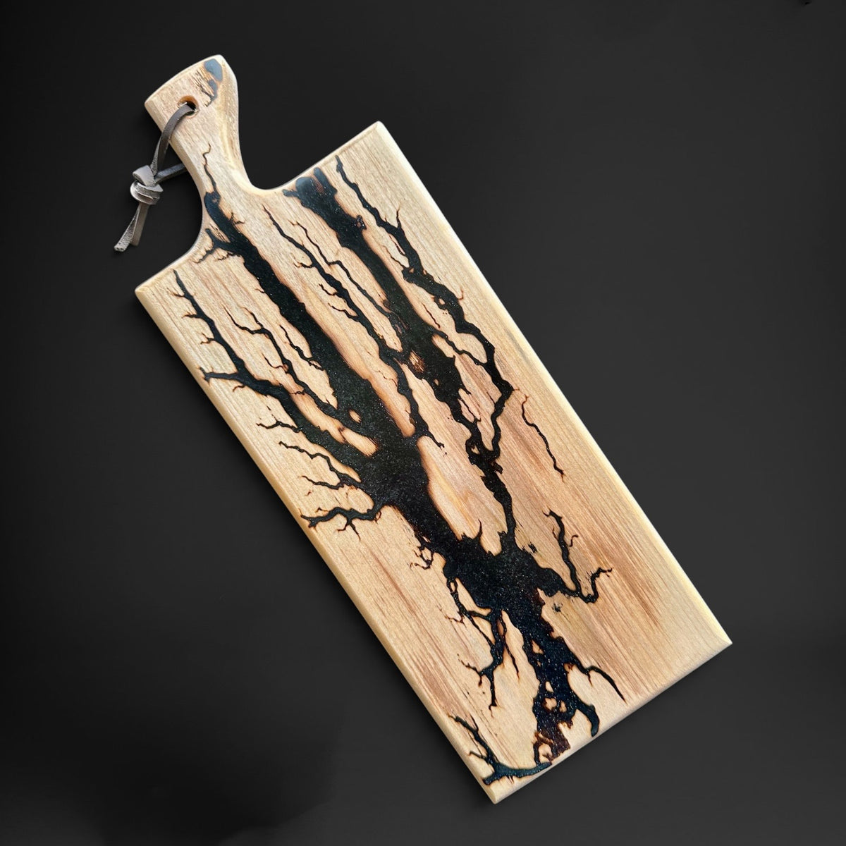 A rectangular wooden cutting board with a natural light wood grain and a dramatic black resin or burn pattern running diagonally across it. It features a built-in handle with a hanging loop at the top, shown on a dark gray background.
