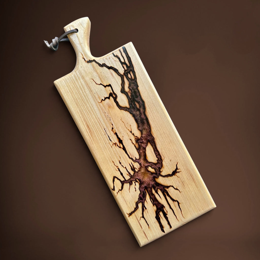 A rectangular wooden cutting board with a natural wood grain finish and a dark brown burnt tree design running down the center. It features a rounded handle and a small leather hanging loop at the top, shown on a solid dark brown background.