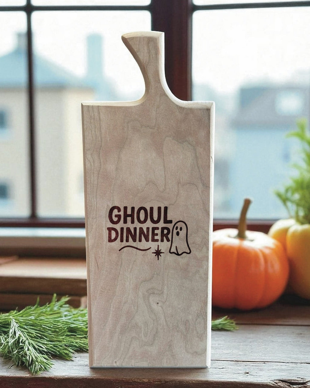 A rectangular wooden cutting board with a handle, featuring the words 'GHOUL DINNER' and a small ghost graphic printed on the front. The board is displayed on a wooden windowsill with autumn decorations, including pumpkins and greenery, in the background.