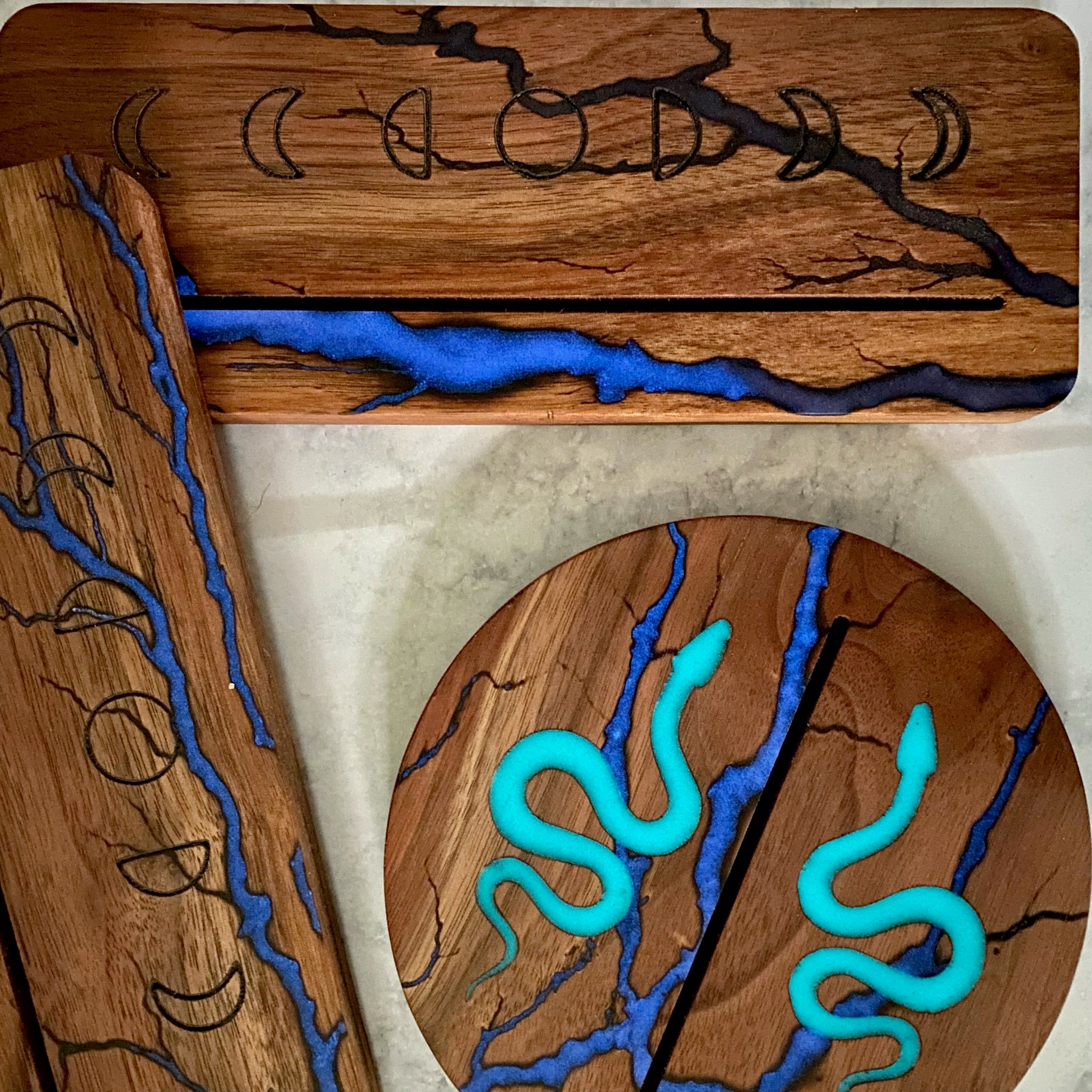 Wooden decorative items with blue resin river details, including a rectangular board with moon phases, a cylindrical coaster with turquoise snakes, and a matching rectangular piece, arranged on a light stone surface.
