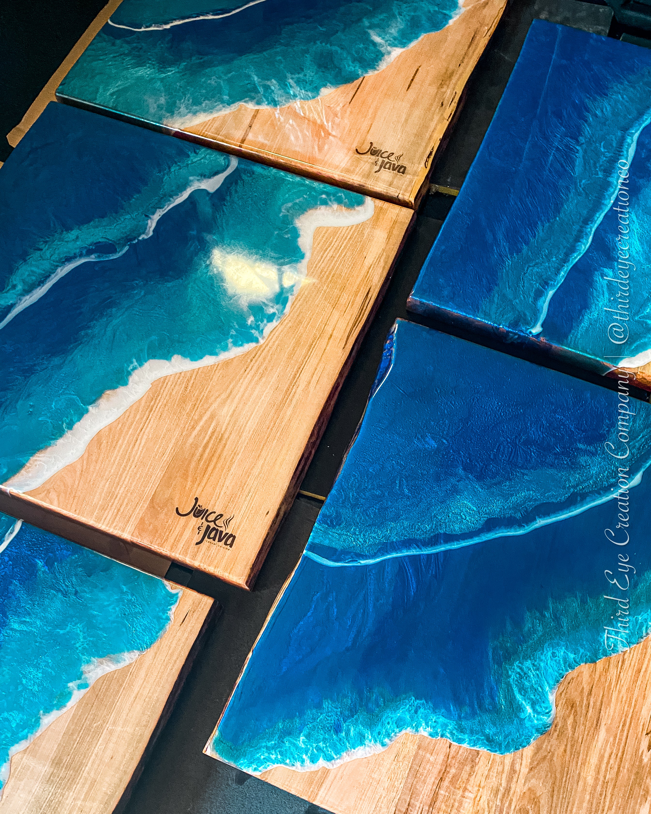 Multiple rectangular resin and wood ocean tabletops arranged on a dark surface, featuring turquoise, blue, white surf, and tan live edge wood grain with the engraved text ‘Juice & Java’.