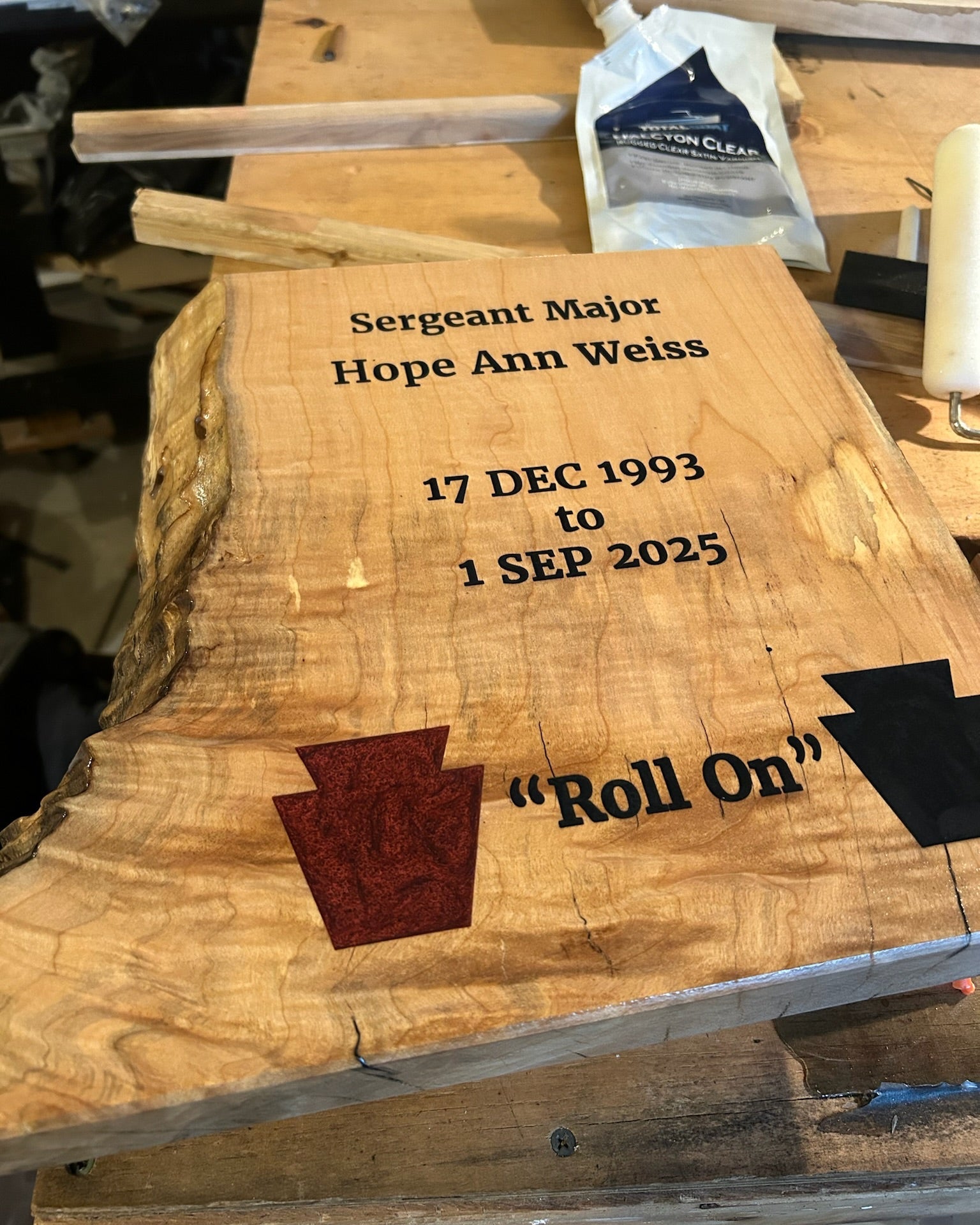 A live-edge wooden maple with engraved text reading 'Sergeant Major Hope Ann Weiss 17 Dec 1993 to 1 Sep 2025' and 'Roll On,' plus two keystone inlays in red and black, photographed on a workbench.