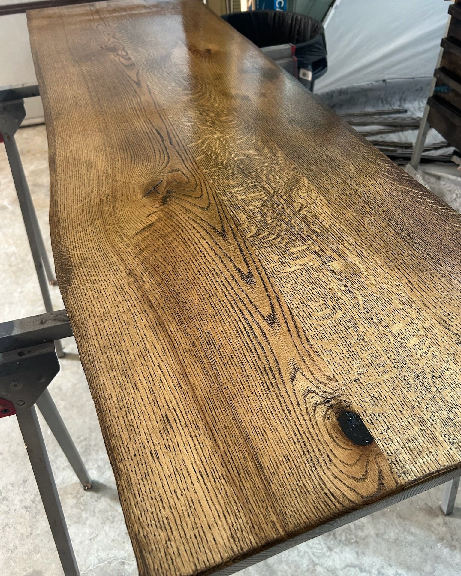 A rustic rectangular wooden live edge table top with a rich brown finish, showcasing prominent natural wood grain, knots, and a live edge, supported by metal legs in a workshop setting.