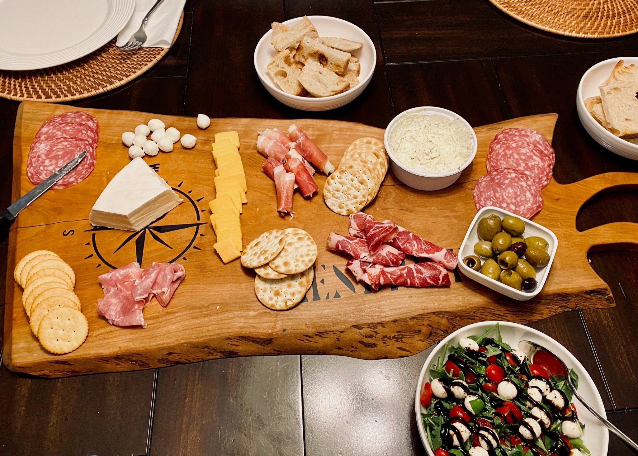 A large rustic maple wooden charcuterie board arranged on a dark wooden table with an assortment of sliced meats, crackers, cheese wedges, olives, and side bowls including a salad and bread.