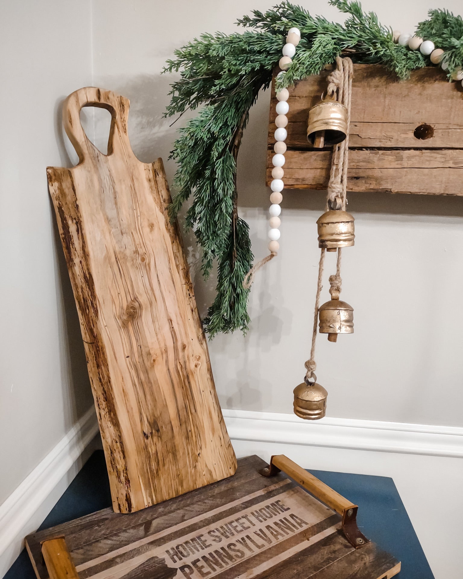 A rustic maple wooden serving board with a live edge and loop handle leans against a wall beside a decorative wooden shelf with green garland, white bead garland, and hanging brass bells. A smaller wooden tray sits on a blue surface in the foreground.