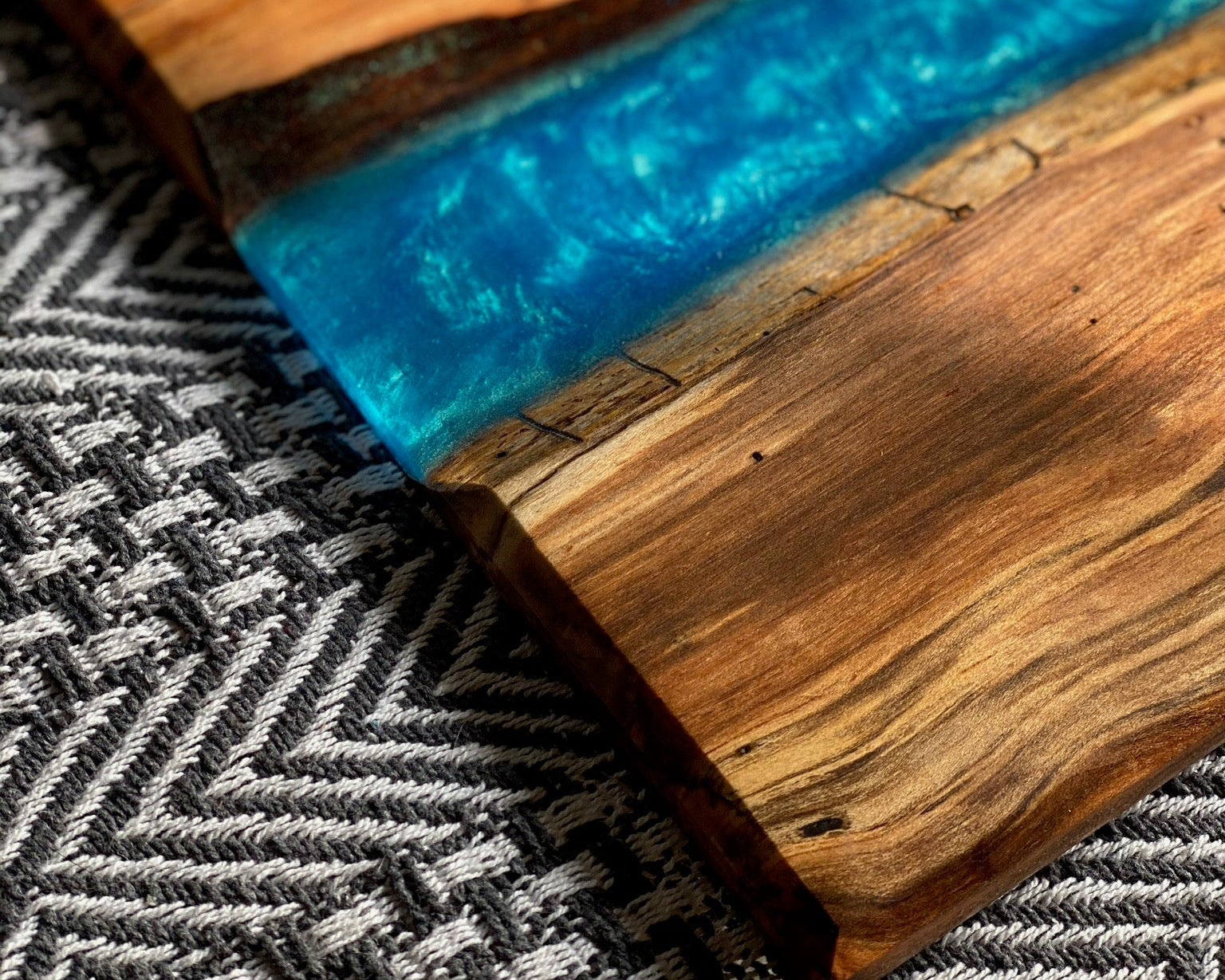 A close-up of a rectangular maple serving  board with a vibrant blue resin river running through the center, resting on a textured gray and white woven rug.