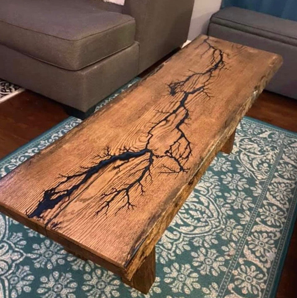 An oak coffee table with a live edge and a prominent fractal burned blue branch-like inlay running diagonally across the top, shown in a living room setting on a teal patterned rug.