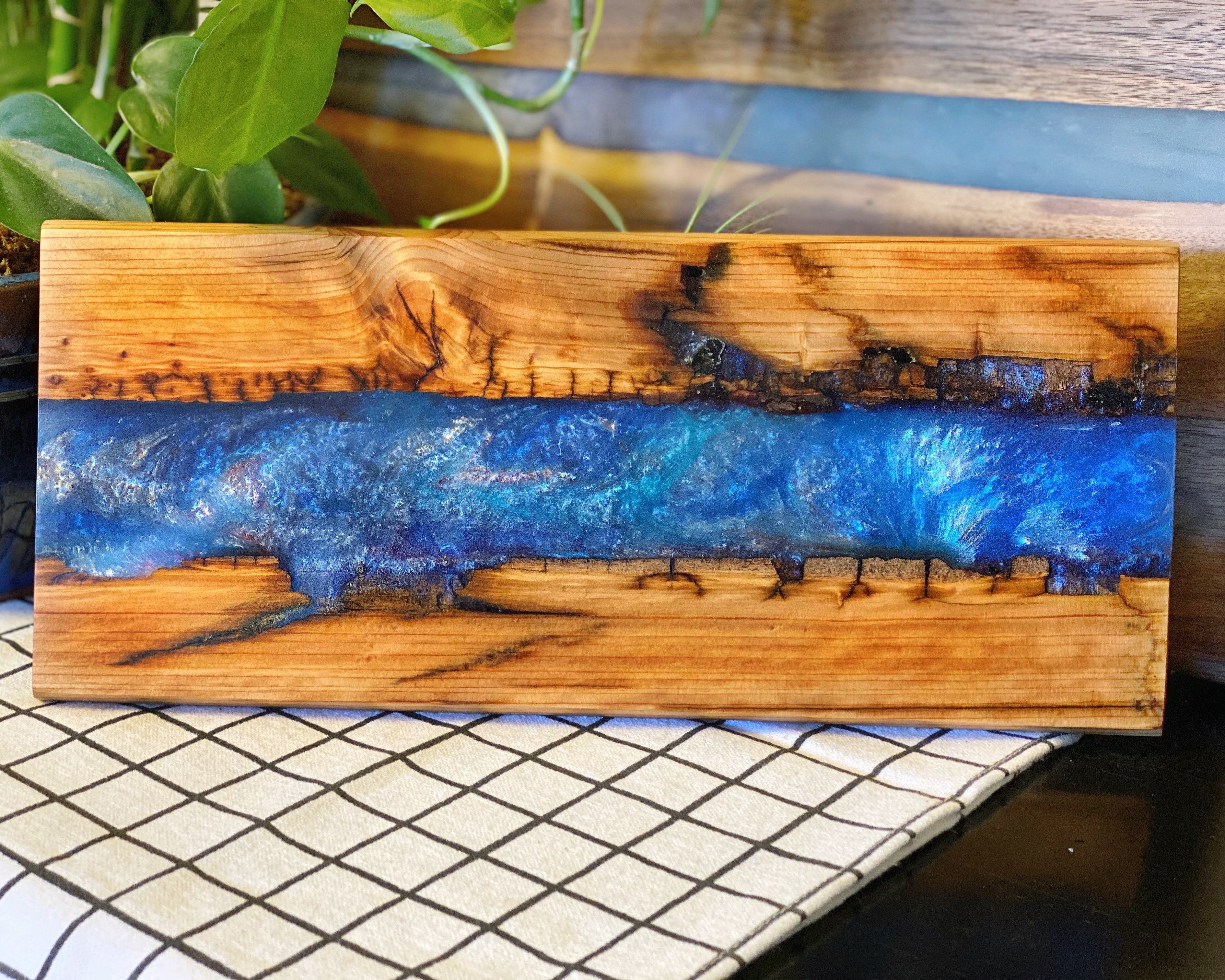 A rectangular wooden serving board with a vibrant blue resin river design running horizontally through the center, set against a rustic wood background with a potted plant and a black-and-white checkered surface.