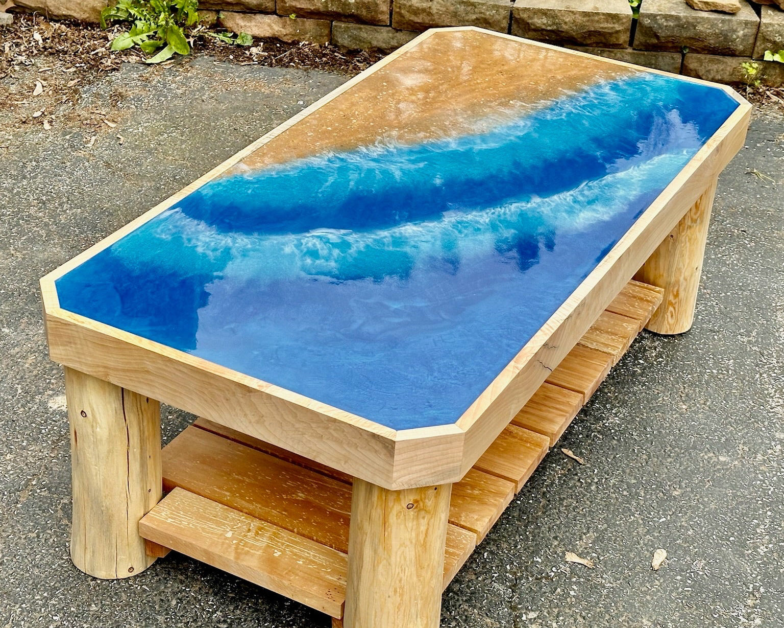 A wooden coffee table with a wave-themed resin top, featuring a natural wood frame and legs, set on an outdoor paved surface.