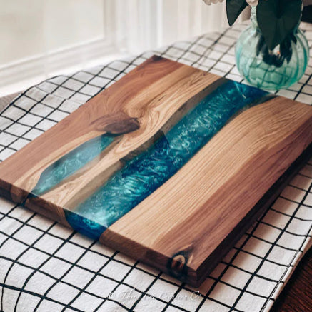 A rectangular cedar cutting board with a live edge and a blue resin river design, displayed on a black and white checkered cloth.