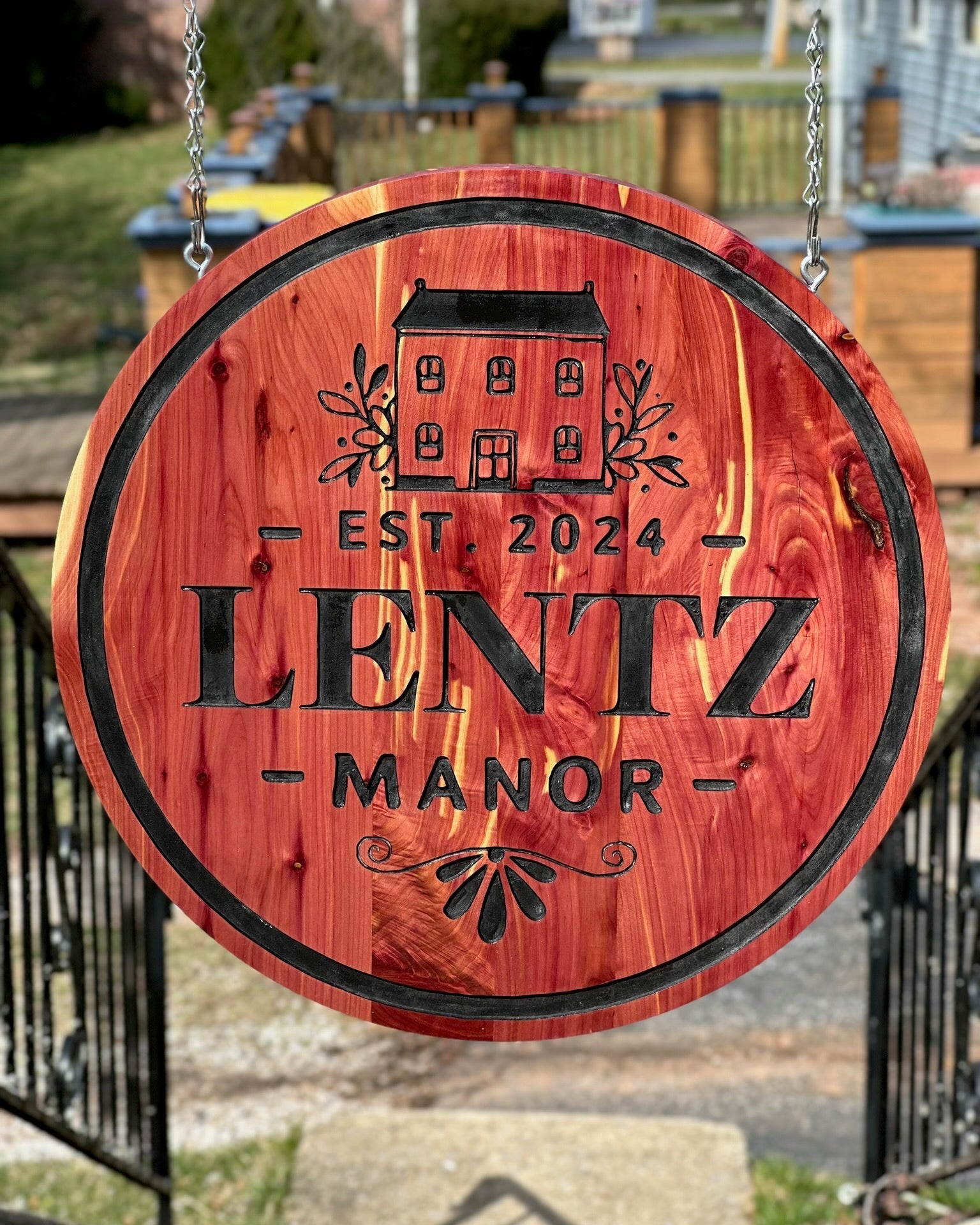 Round wooden hanging sign in cedar and black engraved text reading 'LENTZ MANOR' and 'EST. 2024', suspended by chains outdoors in front of a gate and house.