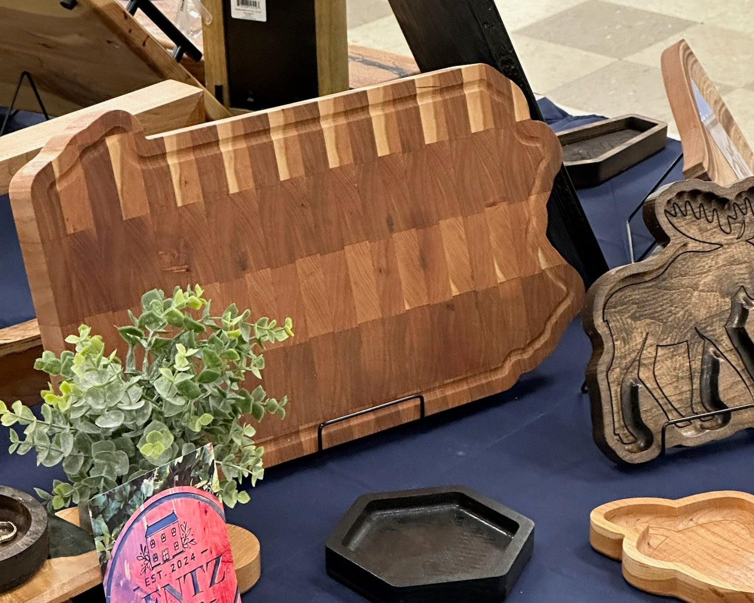 A wooden PA state shaped cutting board displayed on a blue tablecloth at a craft fair, alongside other handmade wooden items including a carved moose plaque, a small black hexagonal tray, and a wooden acorn dish.