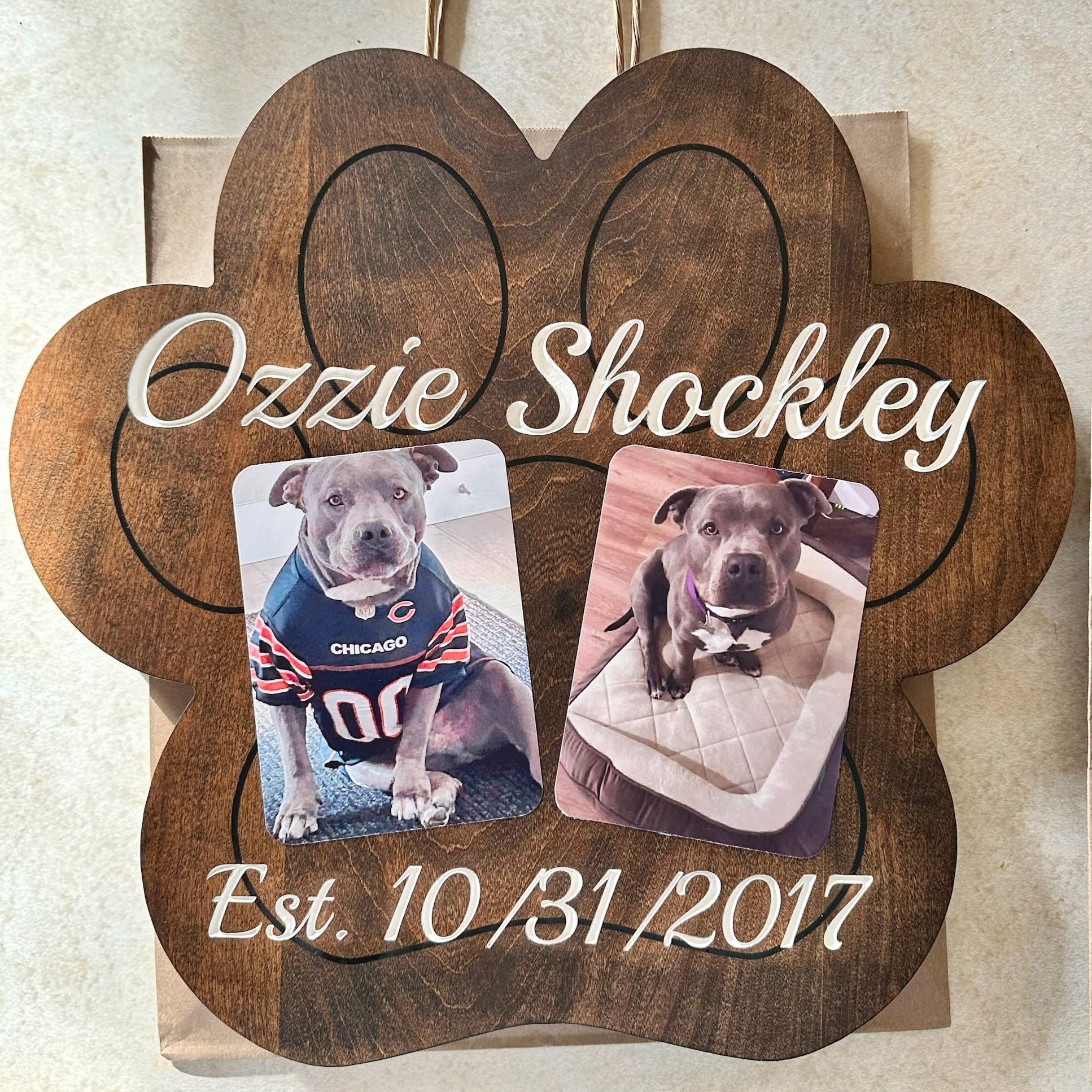 A custom wooden paw-shaped memorial plaque featuring the engraved text 'Ozzie Shockley' and 'Est. 10/31/2017' with two custom pet photos of a pitbull-type dog, displayed on a light beige surface.